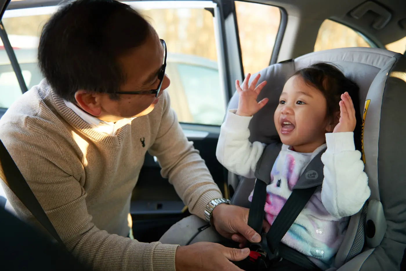 Ett småbarn sitter i en bakåtvänd bilbarnstol och barnets pappa håller på att spänna fast säkerhetsbältet.