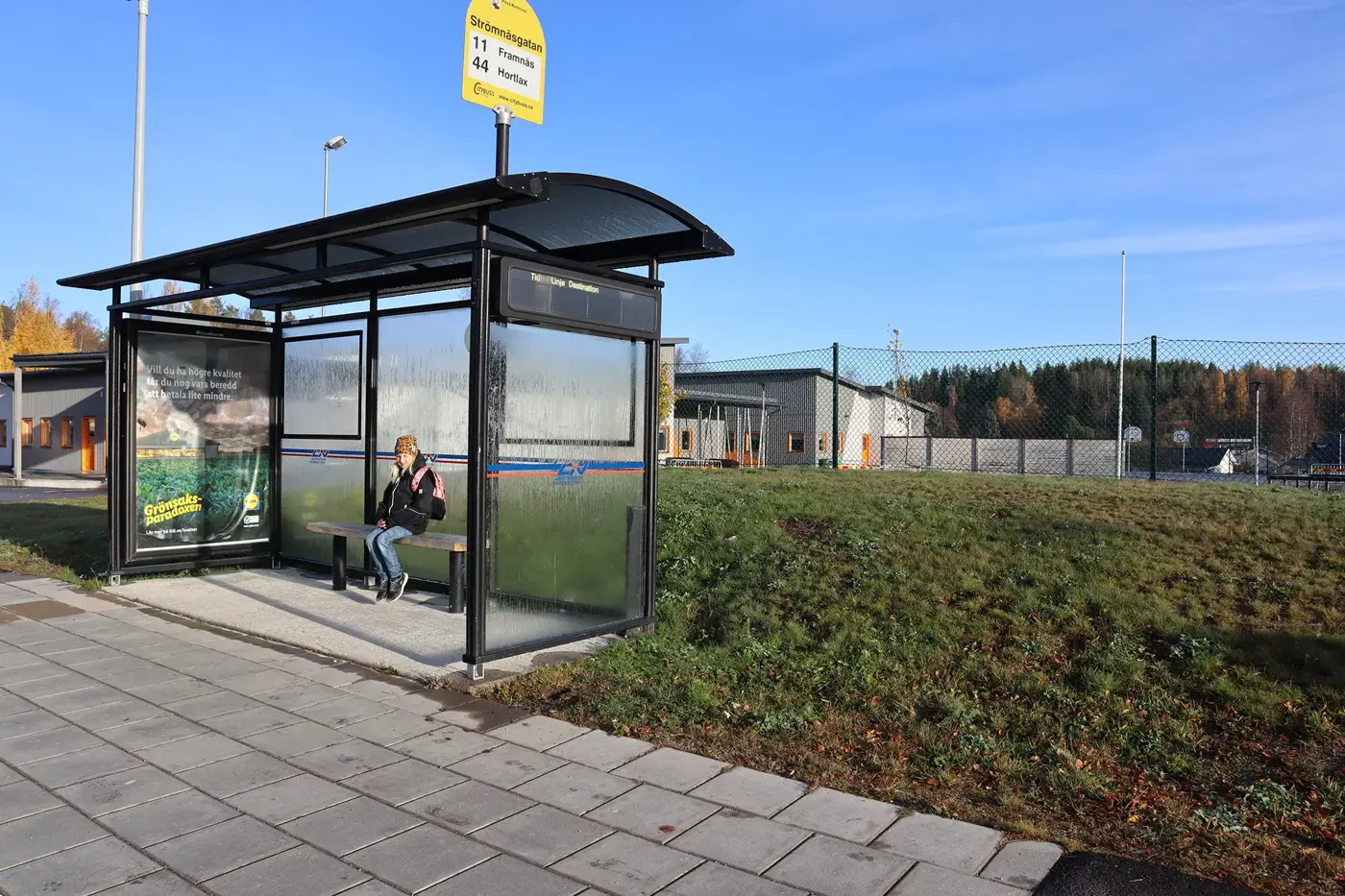 En busshållplats i glas där en flicka sitter på en bänk