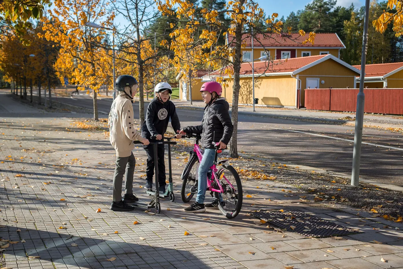 Tre barn med hjälm på huvudet står och pratar på en gång-och cykelbana. Två står med varsin kickbike och den tredje med en cykel.