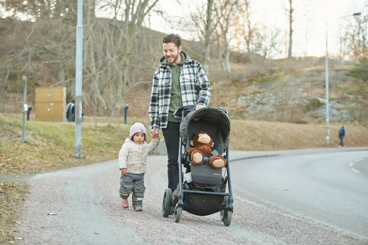 En pappa och hans lilla barn promenerar och kör en vagn där det sitter ett gosedjur.