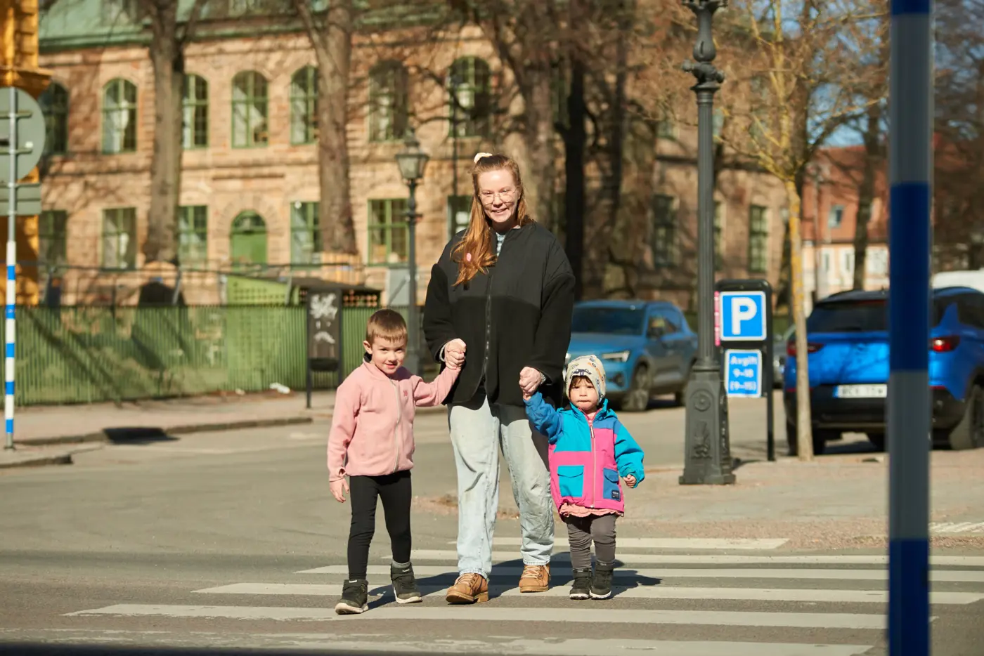 En förälder håller två barn i handen och går över ett övergångsställe. Bilden är tagen framifrån.