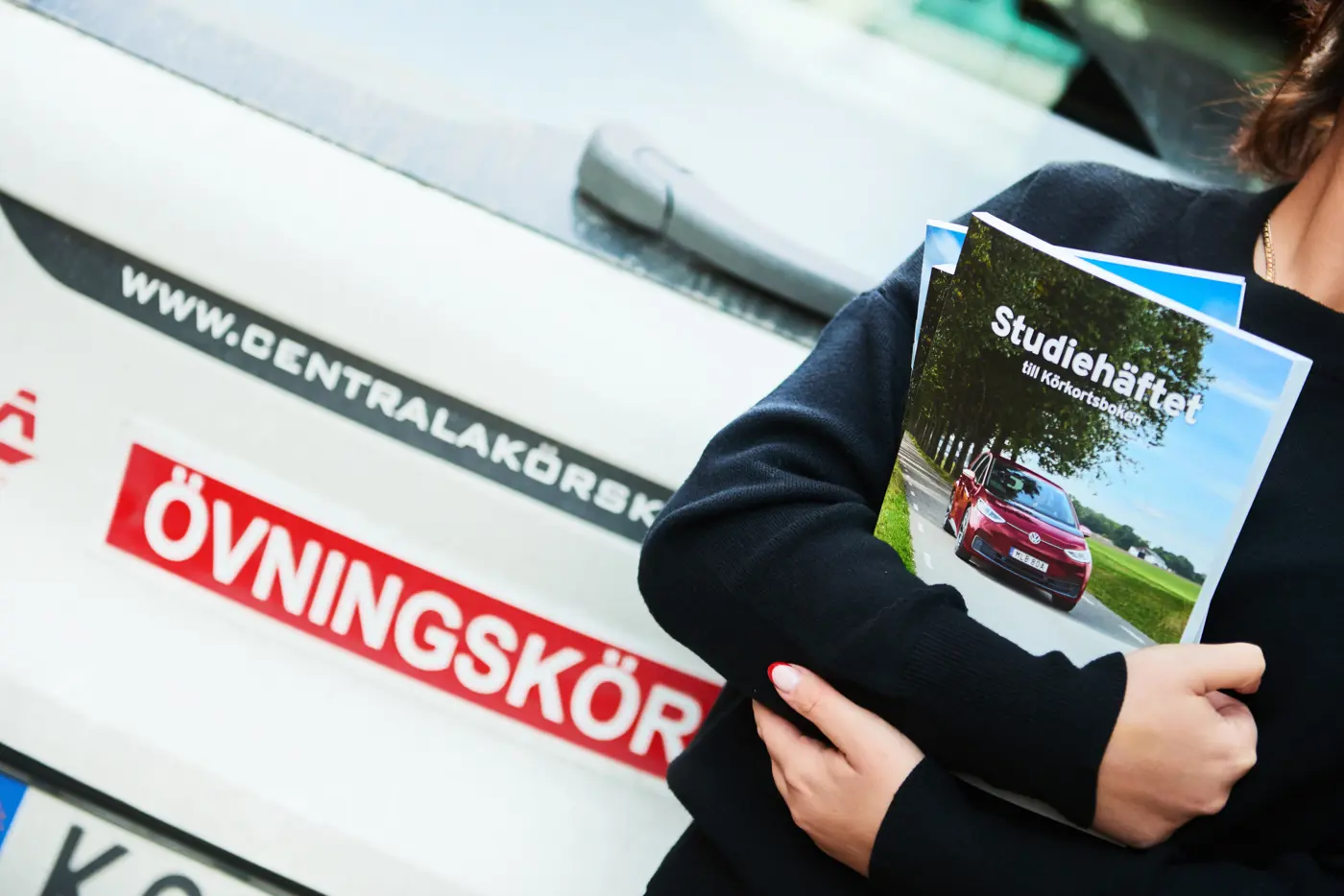 Person som håller i läroböcker om bilkörning framför en bil med skylt för övningskörning