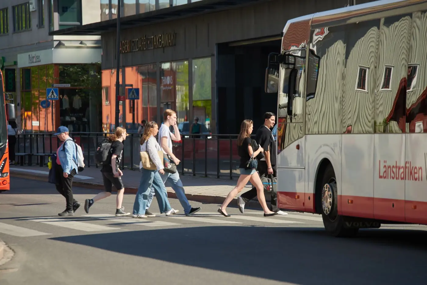 Människor i olika åldrar går över ett övergångsställe i stadsmiljö där två bussar står och väntar på varsin sida.